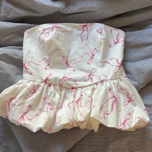 Vit tubtopp med rosa rosett-mönster - Supersöt vit tubtopp med volang nertill och rosa rosett-mönster. Toppen har en smockad del baktill för extra komfort och sitter snyggt. Perfekt för dig som vill ha en lekfull och trendig look!