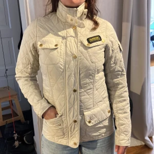 Beige quiltad jacka från Barbour International - Säljer en beige quiltad jacka från Barbour International med guldiga knappar och dragkedja. Jackan har hög krage, flera fickor och en klassisk Barbour-logga på bröstet. Perfekt för vår och höst, snygg och tidlös design.
