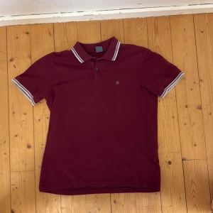 Vinröd pikétröja från Calvin Klein - Säljer en vinröd pikétröja från Calvin Klein med vit- och rödrandiga detaljer på krage och ärmslut. Klassisk polo med knappslå och broderad ck-logga på bröstet. Perfekt för en stilren och avslappnad look.