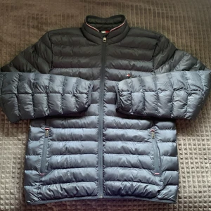 Svart pufferjacka från Tommy Hilfiger - Säljer en marinblå jacka från Tommy Hilfiger med diskreta logodetaljer och dragkedja framtill. Jackan har två fickor med dragkedja och hög krage. Perfekt till vår, sommar och höst. Den är testad en gång så den är i princip helt ny. Säljer den då den är för stor.