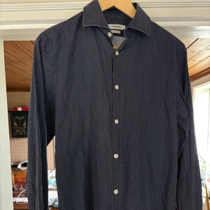 Mörkblå slim fit skjorta från J.Lindeberg - Snygg mörkblå skjorta från J.Lindeberg i slim fit-modell. Skjortan har ett diskret vitt prickigt mönster, klassisk krage och knäppning framtill. Tillverkad i 100% bomull för en skön känsla och stilren look.