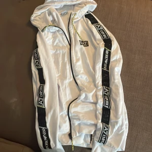 Vit hoodie från Emporio Armani EA7 med svarta detaljer - Säljer en vit hoodie från Emporio Armani EA7 med svarta band och loggor längs ärmarna och axlarna. Jackan har huva, dragkedja och gula detaljer på snöret. Perfekt för en sportig och trendig look.