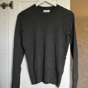 Mörkgrå slätstickad tröja från Holly & Whyte - Säljer en stilren mörkgrå slätstickad tröja från Holly & Whyte. Tröjan har rund halsringning, långa ärmar och ribbade muddar. Den har guldknappar på ärmsluten. Perfekt basplagg för garderoben och passar till många olika outfits. 