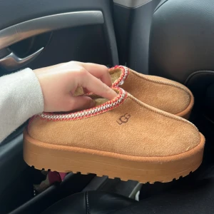Bruna slip-in tofflor från UGG - Mysiga bruna slip-in tofflor från UGG med mjukt foder och dekorativ rödvit kant runt öppningen. Skorna har en rejäl platt sula och är perfekta för kalla dagar. Utsidan är i mocka och insidan är fodrad med pälsimitation. Liknar Ugg (inte äkta) 