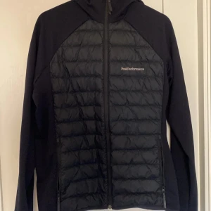Svart hybridjacka från Peak Performance - Svart hybridjacka från Peak Performance med quiltad front och rygg samt huva. Jackan har dragkedja framtill och två sidofickor med dragkedja. Perfekt för lager-på-lager och snygg sportig look. Köpte här på Plick 