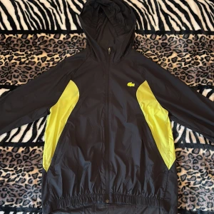 Svart och gul vindjacka från Lacoste - Snygg svart vindjacka från Lacoste med gula paneler på sidorna och huva. Jackan har dragkedja framtill, elastiska muddar och två fickor med dragkedja. Perfekt för en sportig look och skyddar mot vinden.