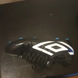 Svarta fotbollsskor från Umbro - Säljer ett par svarta fotbollsskor från Umbro med vit logga på sidan och blå dobbar undertill. Skorna har snörning och är tillverkade i syntetmaterial. Perfekta för dig som vill ha stilrena och sportiga skor på planen.