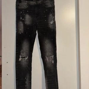 Svarta jeans med slitningar från Supply & Demand - Snygga svarta jeans från Supply & Demand med slitningar på knäna och små vita färgstänk. Jeansen har fem fickor och en klassisk jeansknäppning. Passar perfekt till en trendig streetwear-look.