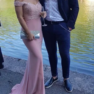 Ljusrosa långklänning med spetsdetaljer - Elegant ljusrosa långklänning med offshoulder och vackra spetsdetaljer upptill. Klänningen har figurnära passform och en lätt utsvängning. Perfekt för bal eller festliga tillfällen. Den har även en sydd slits på ena låret.  Passar storlek XS-S.                                                   Ordinarie pris på klänningen är 5000:-  