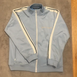Ljusblå track jacket från Palm Angels - Säljer en ljusblå track jacket från Palm Angels med vita och svarta ränder längs ärmarna. Jackan har dragkedja framtill, ståkrage och ribbade muddar. Perfekt för en sportig och avslappnad stil.