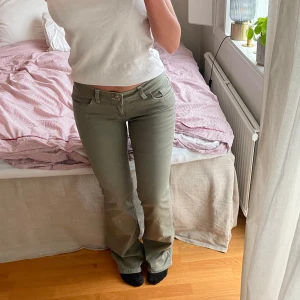 Olivgröna jeansbyxor från Nelly - Snygga olivgröna jeansbyxor från Nelly med vita kontrastsömmar och dekorativ broderad bakficka. Byxorna har låg midja, bootcut-modell och dubbla knappar framtill🌺