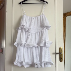 Vit volangklänning från NA-KD - Superfin vit klänning från NA-KD med tunna axelband och flera lager volanger. Klänningen har en plisserad struktur och är perfekt för sommarens alla tillfällen. Säljs med originaltag kvar. Även perfekt som studentklänning. 