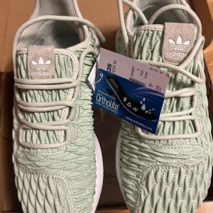 Adidas Tubular Shadow W Linen Green - Snygga Adidas Tubular Shadow W i färgen Linen Green. Skorna har en unik texturerad ovandel med snörning och vit platt sula. Perfekta sneakers för dig som vill sticka ut med stilren design och bekväm passform.