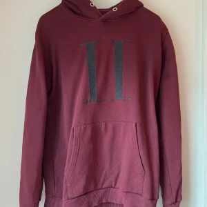 Vinröd hoodie från Les Deux - Nypris 1299 kr. Vinröd hoodie från Les Deux i storlek XL. Tröjan har en stor ficka framtill, luva och tryckt svart/grå logga på bröstet. Tillverkad i mjuk bomullsblandning med 80% bomull och 20% polyester.