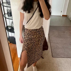 Leopardmönstrad kjol med slits - Säljer en snygg leopardmönstrad kjol i midi-längd med slits på sidan. Kjolen har en beige bas med svarta och bruna leopardfläckar. Passformen är normal och materialet känns mjukt och följsamt, troligtvis polyester. Perfekt statement-plagg för dig som gillar djurmönster.