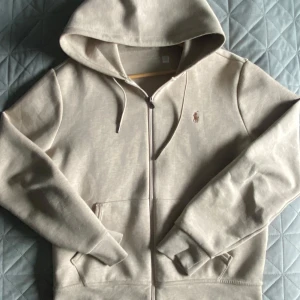 Beige hoodie från Ralph Lauren - Snygg beige hoodie från Ralph Lauren med dragkedja och huva. Tröjan har broderad logga på bröstet och praktiska fickor framtill. Perfekt för en avslappnad och stilren look.