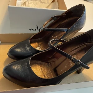Svarta Mary Jane-klackar från Nilson - Säljer ett par klassiska svarta Mary Jane-klackar från Nilson med rund tå och justerbart spänne över foten. Har en elegant, tidlös design som passar till många olika outfits.