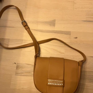 Ljusbrun axelväska med dekorativa detaljer - Säljer en ljusbrun axelväska i syntetmaterial med justerbar rem. Väskan har ett dekorativt spänne framtill med små pärlor och ett stilrent, enkelt bakstycke. Perfekt för att bära det viktigaste i vardagen. Pärlorna går att ta bort. 