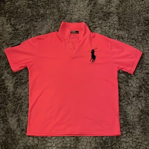Rosa pikétröja från Polo Ralph Lauren - Säljer en klassisk rosa pikétröja från Polo Ralph Lauren med stor svart logga broderad på bröstet. Tröjan har korta ärmar, krage och knappar vid halsen. Perfekt för en sportig och stilren look.