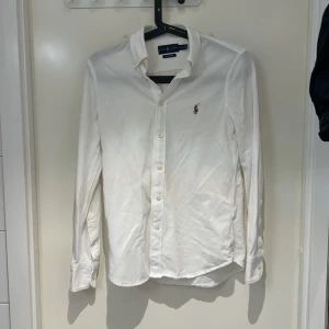 Vit skjorta från Ralph Lauren - Klassisk vit skjorta från Ralph Lauren med broderad logga på bröstet. Pris går att diskutera skjortan har lång ärm, knappar framtill och är tillverkad i bomull. Perfekt för en stilren och tidlös look.