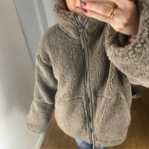 Beige teddyjacka med dragkedja - Mysig beige teddyjacka med hög krage och dragkedja framtill. Jackan har två stora fickor och en avslappnad passform, perfekt för kyliga dagar. Snygg och trendig att bära till vardags.