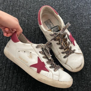 Golden Goose sneakers med röd stjärna - Golden Goose sneakers i vitt skinn med röd stjärna på sidan och röd häl. Skorna har leopardmönstrade snören och guldiga detaljer på plösen. Klassisk rund tå och platt sula. Perfekta för dig som gillar en unik och trendig stil.