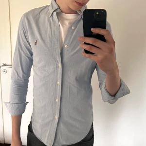 Ljusblå skjorta från Ralph Lauren - Säljer en klassisk ljusblå skjorta från Ralph Lauren med broderad logga på bröstet. Skjortan har lång ärm, knappar framtill och en normal passform. Perfekt till jeans eller chinos. Använd men bra skick!