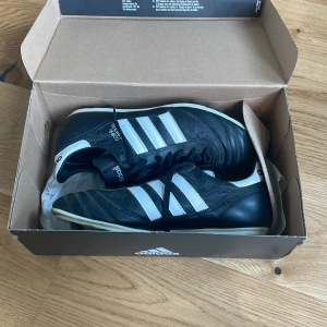 Adidas Copa Mundial fotbollsskor - Klassiska svarta Adidas Copa Mundial fotbollsskor med vita ränder och snörning. Skorna har rund tå, platt sula med fasta dobbar och är tillverkade i skinn. Perfekta för fotboll på gräsplan. Har original kartong. Perfekta för gräs! Nypris 1200!