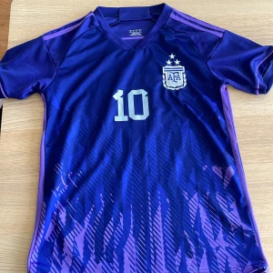 Argentina Messi fotbollströja  - Säljer en lila Argentina fotbollströja med nummer 10 och Messi på ryggen. Tröjan har korta ärmar, AFA-emblem på bröstet och ett mönster i olika nyanser av lila och blått. Perfekt för fotbollsfans!
