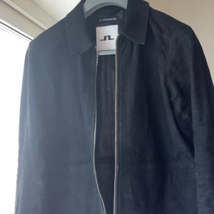 Svart mockajacka från J.Lindeberg - Stilren svart mockajacka från J.Lindeberg med klassisk krage och dragkedja framtill. Jackan har en minimalistisk design och är perfekt för dig som gillar en clean look. Tunn så passar utmärkt till sommaren. 100% mocka. Nypris: 5200kr. M men passar S också. 