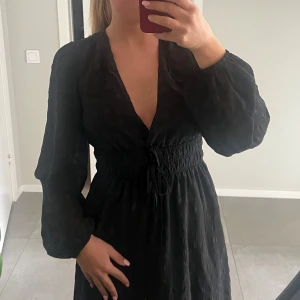 Svart långklänning med v-ringning och knyt i midjan - Svart långklänning med diskret rutmönster, djup v-ringning och långa ärmar. Klänningen har markerad midja med knytband och lätt transparenta ärmar. Perfekt för dig som vill ha en elegant och stilren look.