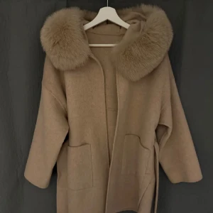 Beige kappa med fuskpäls från Instagram  - Säljer en beige kappa med stor huva och bred fuskpäls runt kragen. Jackan har två stora fickor framtill och ett bälte i midjan för att knyta. Perfekt för kyliga dagar och ger en lyxig känsla.
