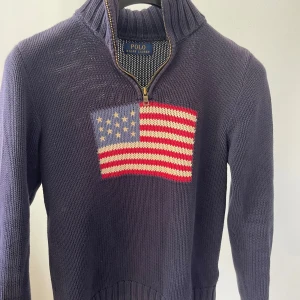 Lila stickad tröja med dragkedja från Polo Ralph Lauren - Säljer en lila stickad tröja från Polo Ralph Lauren med halv dragkedja och hög krage. På bröstet finns en stor amerikansk flagga i rött, vitt och blått. Tröjan har långa ärmar och ribbade muddar vid ärmslut och nederkant. Passar: XS/S