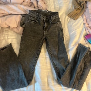 Grå jeans med råa benslut - Säljer ett par grå jeans med hög midja och råa, fransiga benslut. Jeansen har klassisk femficksdesign och dragkedja med knapp framtill. Perfekta för en avslappnad och trendig look.