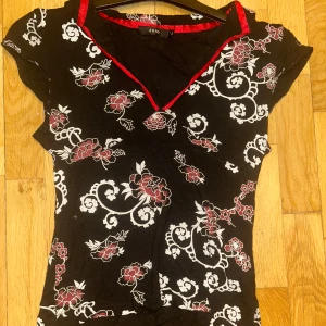 Svart blommig topp med röd kant från Zoio - Säljer en svart topp med vita och röda blommönster samt dekorativ röd kant vid v-ringningen. Toppen har korta ärmar och figurnära passform. Perfekt för dig som gillar unika detaljer och mönster!