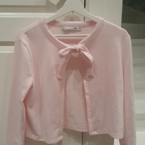 Ljusrosa kofta med rosett - Supersöt ljusrosa kofta med knytrosett framtill. Mjuk och skön, perfekt att slänga över en topp. Långärmad och enkel design som passar till mycket. Väldigt skön och sval nu till sena sommar kvällarna.