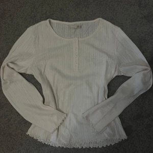 Vit ribbad långärmad topp med knappar - Säljer en söt vit långärmad topp med ribbat mönster och dekorativa små knappar framtill. Toppen har rund halsringning och fina vågiga kanter vid ärmslut och nederkant. Perfekt till lager-på-lager eller som den är.