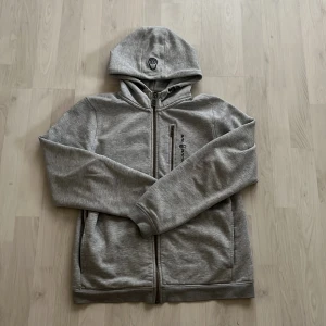 Sail racing zip hoodie - Säljer en grå hoodie från Sail Racing med dragkedja framtill och broderad logga på bröstet. Tröjan har huva och en praktisk bröstficka med dragkedja. Perfekt för en avslappnad stil.