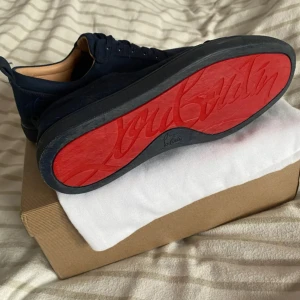 Mörkblå skor från Christian Louboutin - Säljer ett par mörkblå sneakers från Christian Louboutin med klassisk röd sula. Skorna har rund tå, snörning och är tillverkade i mocka. Perfekta för dig som vill ha något stilrent men ändå sticker ut med den ikoniska röda sulan. Har box kvitto påse och allt 