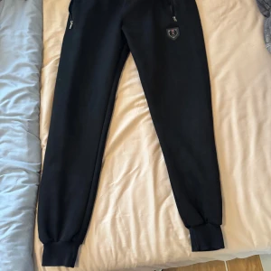 Svarta mjukisbyxor med tröja från XPOOL - Svarta mjukisbyxor från XPOOL med resår i midjan och benslut. Byxorna har dragkedjeförsedda fickor och en broderad XPOOL-logga på ena benet. Perfekta för en avslappnad stil eller till träning.