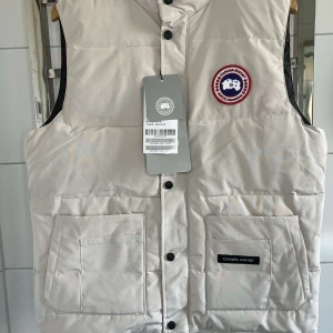 Vit dunväst från Canada Goose - Säljer en snygg vit dunväst från Canada Goose med klassisk logotyp på bröstet. Västen har svarta knappar framtill, två stora fickor och hög krage. Perfekt för lager-på-lager och håller dig varm under kyliga dagar. Helt oanvänd och kommer med kvitto.