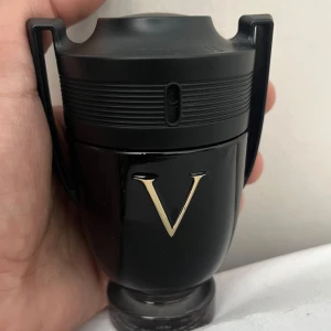 Invictus Victory Eau de Parfum för herr - En lyxig parfymflaska från Rabanne i svart och guld, formad som en pokal med ett stort V på framsidan. Doften har tydliga inslag av vanilj, amber, sötma, aromatiska och kryddiga toner samt fräsch citrus. 93ml kvar av 100ml