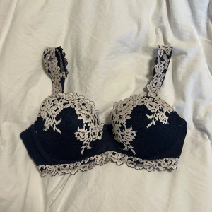 Mörkblå spets bh från intimissimi - Säljer en mörkblå bh med vackra vita spetsdetaljer längs kupor, axelband och nederkant. Bh:n har ett elegant blommönster i spetsen och ger ett lyxigt intryck. Perfekt för dig som vill ha något extra fint under kläderna. Endast testat en gång! Storlek 75C med bygel. 