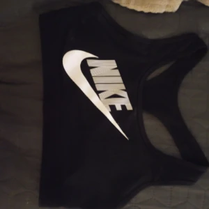Svart sport-bh från Nike - Svart sport-bh från Nike med klassisk vit logga framtill. Modellen har brottarrygg och är tillverkad i ett stretchigt material som passar perfekt för träning.