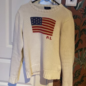 Vit stickad tröja med amerikansk flagga från Polo Ralph Lauren - Vit stickad tröja från Polo Ralph Lauren med amerikansk flagga och RL-broderi på bröstet. Tröjan har rund halsringning och långa ärmar. Klassisk och tidlös design med ribbade muddar vid ärmslut och nederkant.