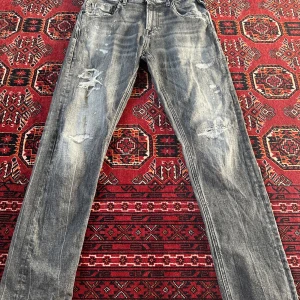 Replay jeans med slitningar - Säljer ett par gråa Replay jeans med slitna detaljer och raka ben. Byxorna har klassisk femficksdesign och tydliga slitningar framtill för en tuff look. Midjan är låg och passformen är normal. Perfekta för dig som gillar en avslappnad stil. Ordinarie pris (2,500kr)