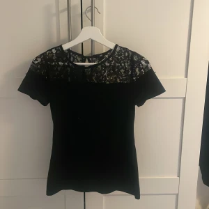 Svart spetsblus från H&M - Säljer en svart blus från H&M med vacker spets upptill och korta ärmar. Blusen har rund halsringning och en liten knapp i nacken. Perfekt för dig som gillar stilrena detaljer och spets.