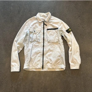 Vit overshirt från Stone Island - Säljer en stilren ljus blå overshirt från Stone Island med svarta detaljer och klassisk patch på ärmen. Jackan har två bröstfickor, dragkedja och knappstängning. Perfekt för lager-på-lager och snygg till många outfits.