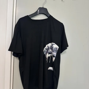 Svart t-shirt med tryck av knytnäve - Cool svart t-shirt med ett unikt tryck av en knytnäve i vitt och grått på framsidan. T-shirten har rund hals och korta ärmar. Perfekt för dig som vill sticka ut med en stilren men ändå uttrycksfull look.