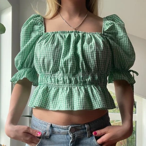 Grön rutig croppad blus med puffärmar - Säljer en söt grön och vit rutig blus med puffiga korta ärmar och volang nertill. Blusen är croppad och har resår i midjan för en bekväm passform. Perfekt för vår och sommar!